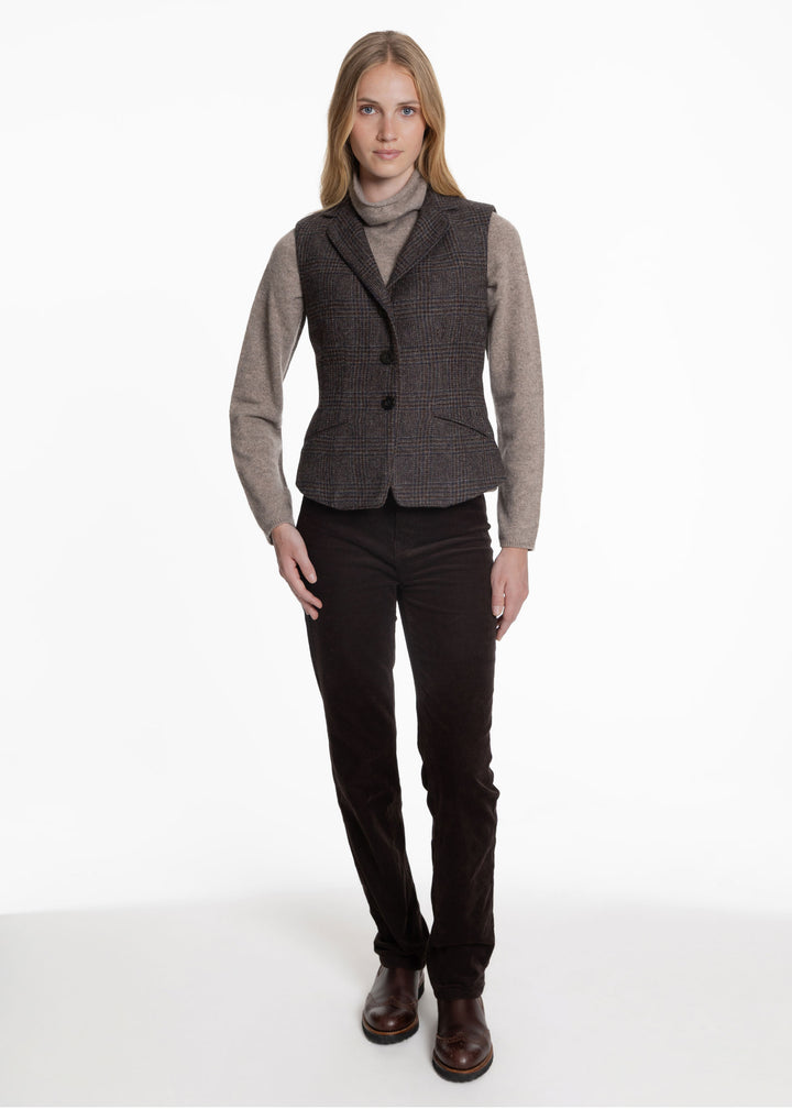 A woman wearing a black an brown check waistcoat over a grey turtleneck sweater. Worn with brown trousers and chelsea boots.