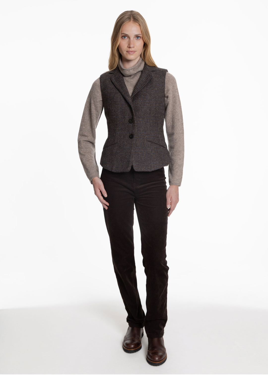 A woman wearing a black an brown check waistcoat over a grey turtleneck sweater. Worn with brown trousers and chelsea boots.