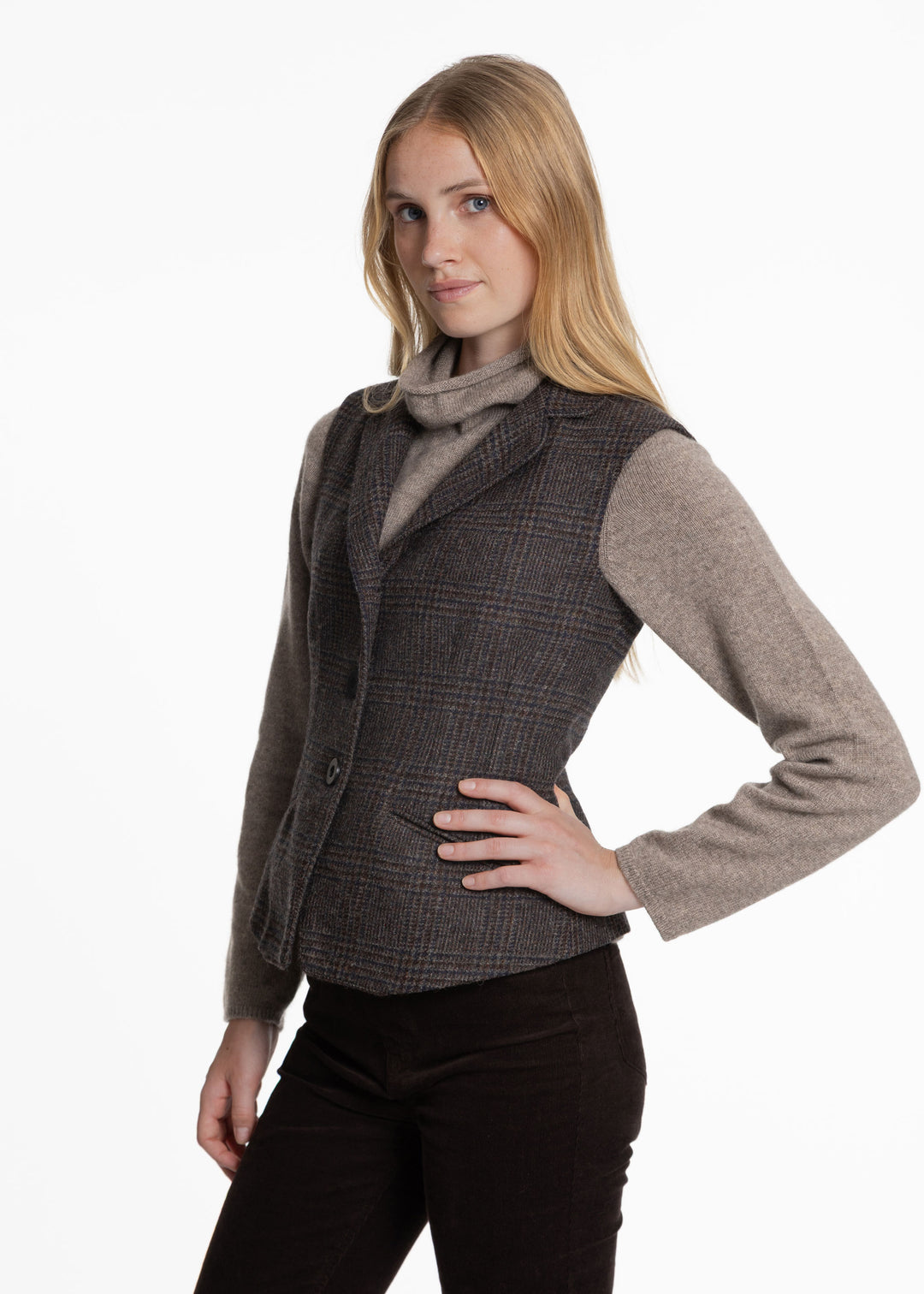 A woman modeling a dark brown and black plaid waistcoat over a taupe turtleneck sweater and dark brown pants, photographed against a plain white studio background.