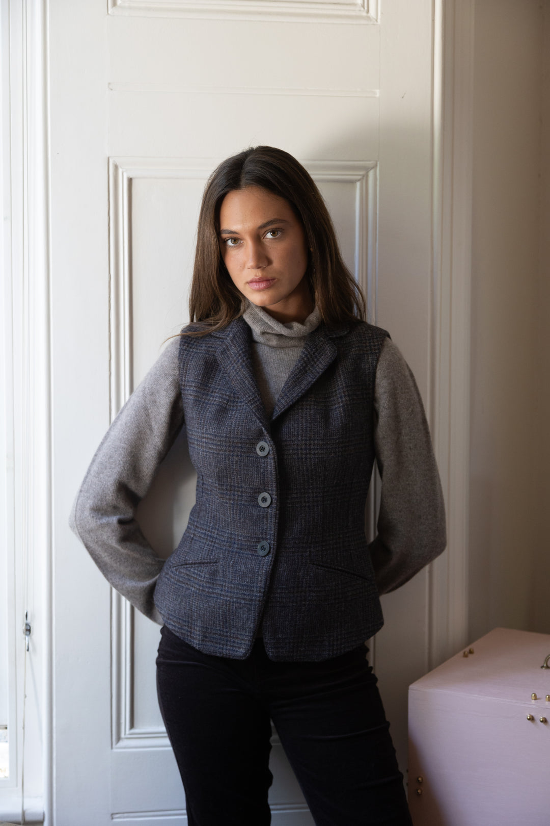 A woman wearing a dark brown and black check waistcoat over a gray turtleneck sweater and black pants, standing in front of a white paneled door.