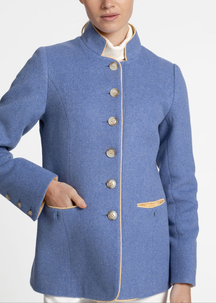A woman wearing a blue wool jacket with a high stand collar, beige buttons, and tan piping details, paired with a cream shirt collar and white trousers, photographed against a light gray studio background.