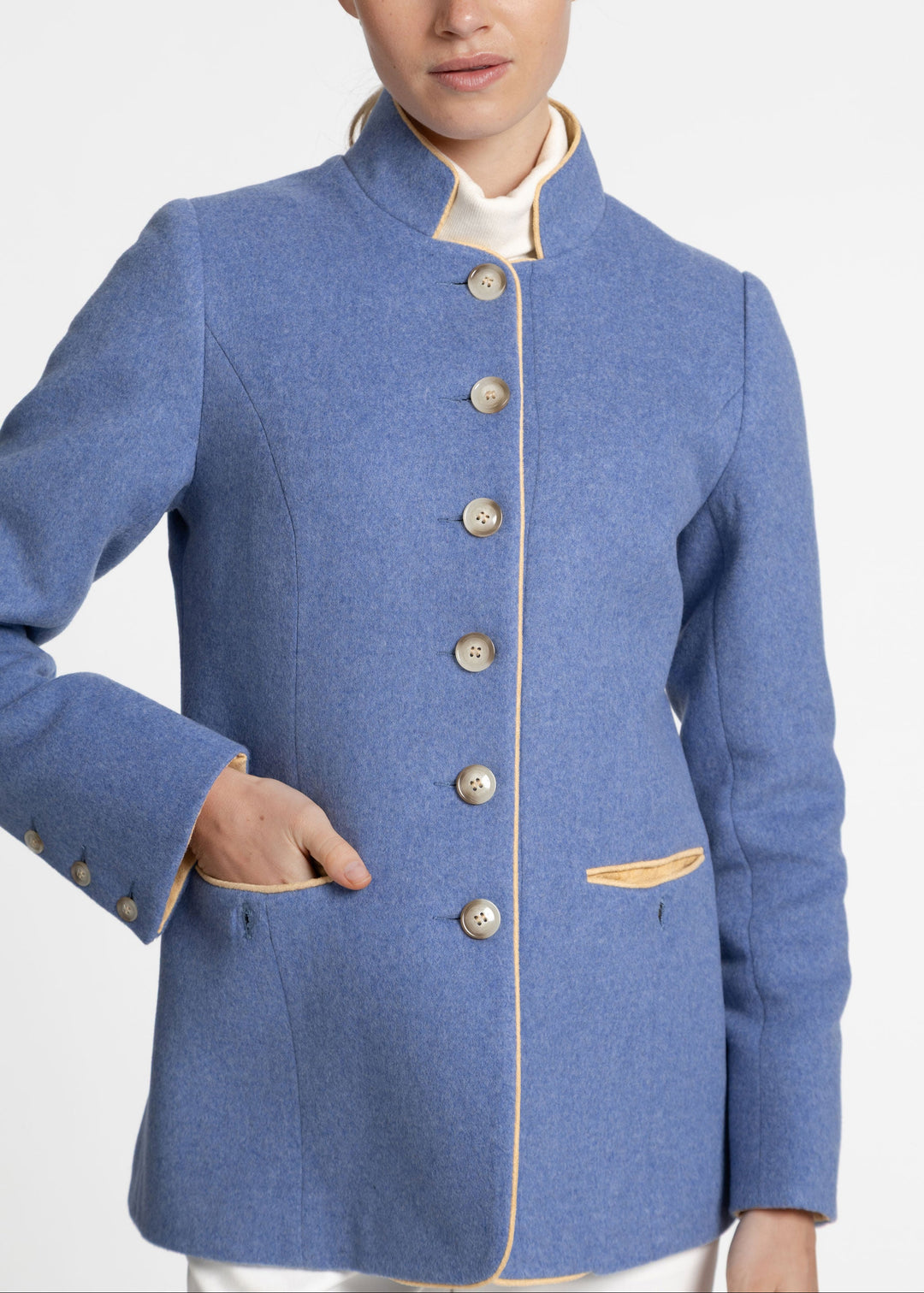 A woman wearing a blue wool jacket with a high stand collar, beige buttons, and tan piping details, paired with a cream shirt collar and white trousers, photographed against a light gray studio background.