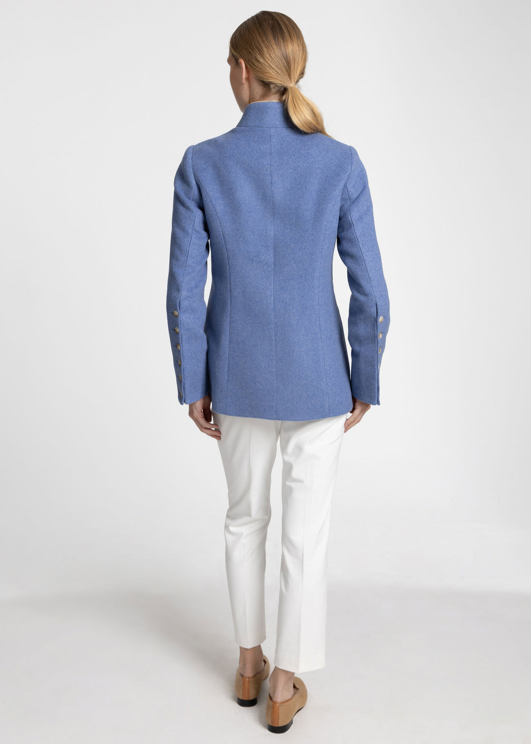 A woman shown from the back wearing a blue structured jacket, white trousers, and tan shoes against a plain light gray studio background.