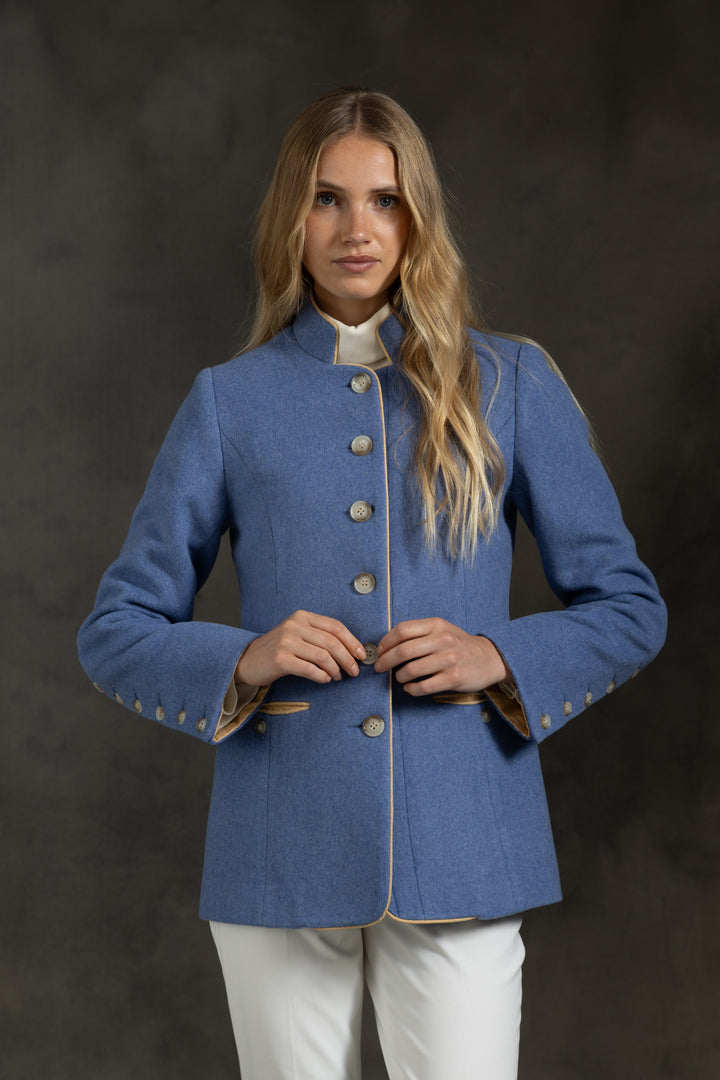 A woman modeling a blue jacket with beige buttons and yellow-beige trim, paired with white trousers, against a dark textured studio background.