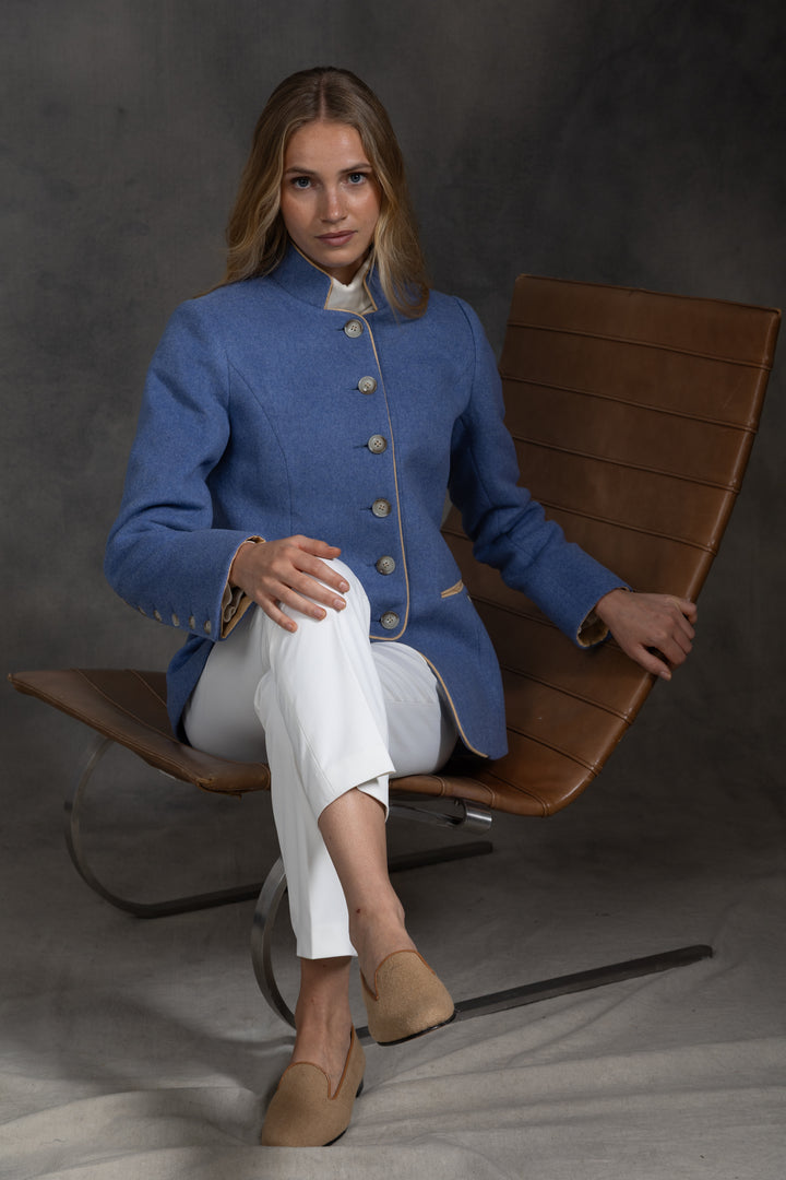 A woman sits in a brown leather chair wearing a blue jacket with beige buttons and white trousers, photographed against a dark gray studio backdrop.