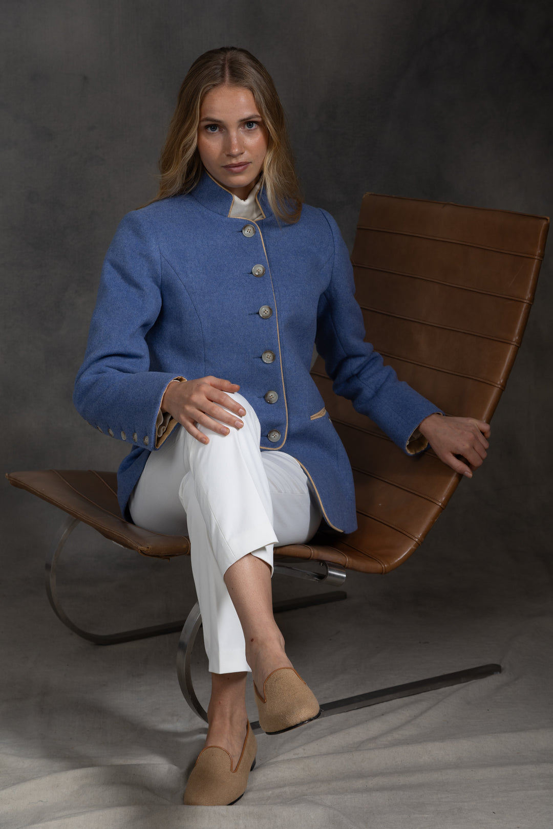 A woman sits in a brown leather chair wearing a blue jacket with beige buttons and white trousers, photographed against a dark gray studio backdrop.
