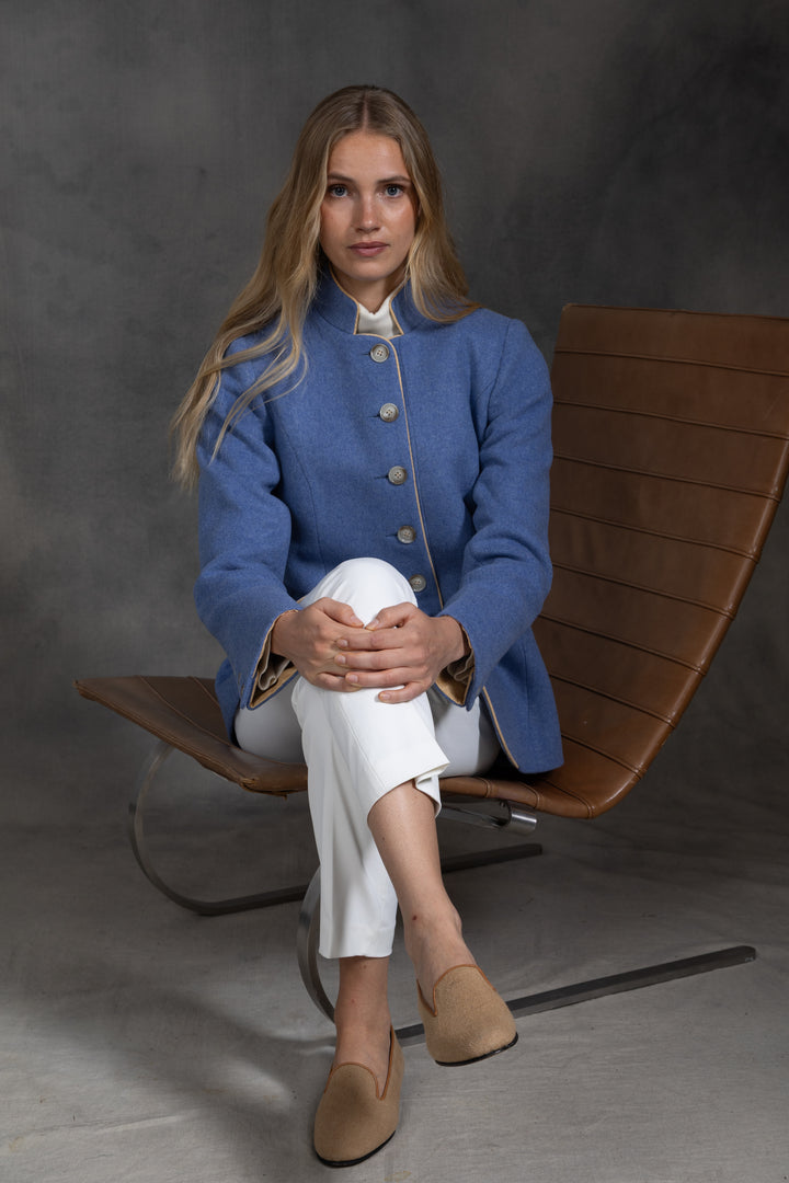 A woman sits on a modern brown leather chair wearing a blue jacket with beige buttons, white trousers, and beige loafers against a dark gray studio background.