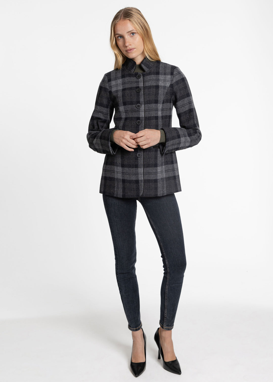 A woman modeling a dark gray and black plaid jacket with dark gray skinny jeans and black pointed-toe heels, standing against a plain white studio background.
