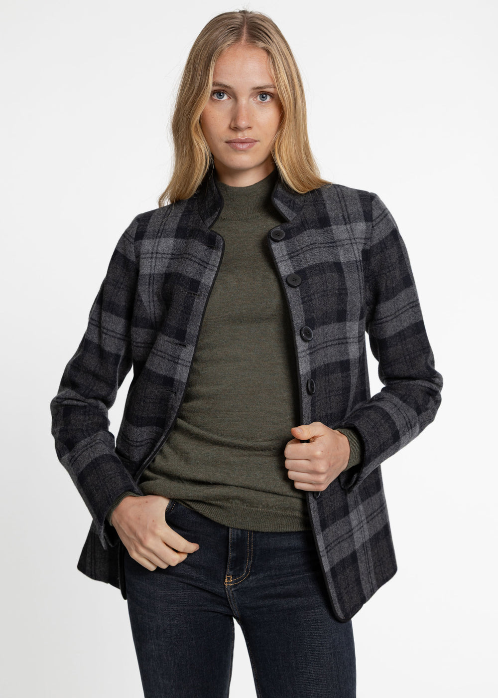 A woman modeling a dark gray and black plaid button-up jacket over an olive green turtleneck sweater and dark blue jeans, photographed against a clean white studio background.