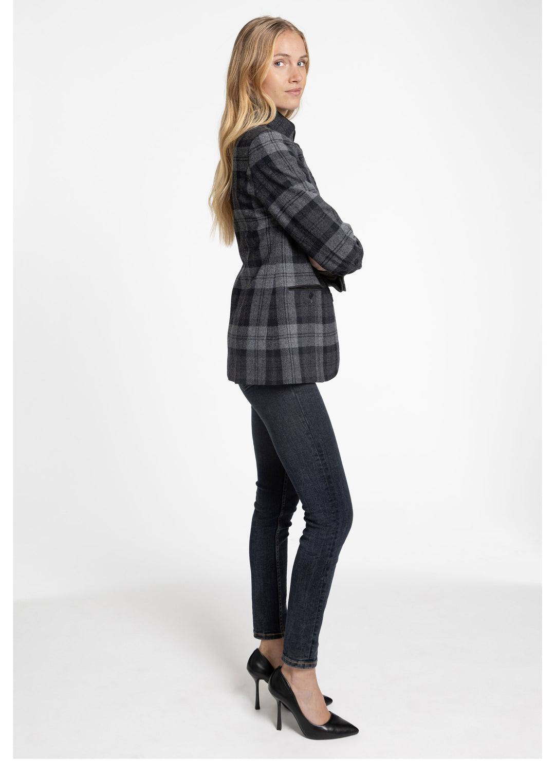 A woman modeling a black and gray plaid jacket, dark skinny jeans, and black high heels against a plain white studio background.