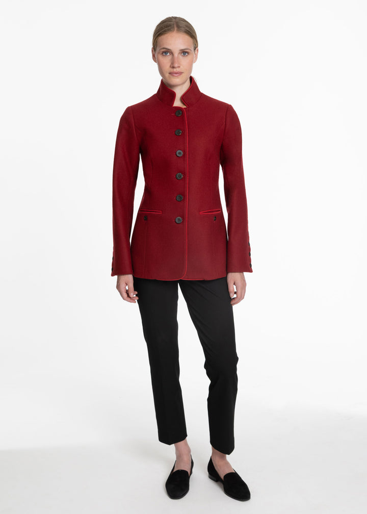 A woman modeling a structured, long-sleeved red jacket with a high stand collar and dark buttons, paired with black trousers and black loafers, against a plain white studio background.