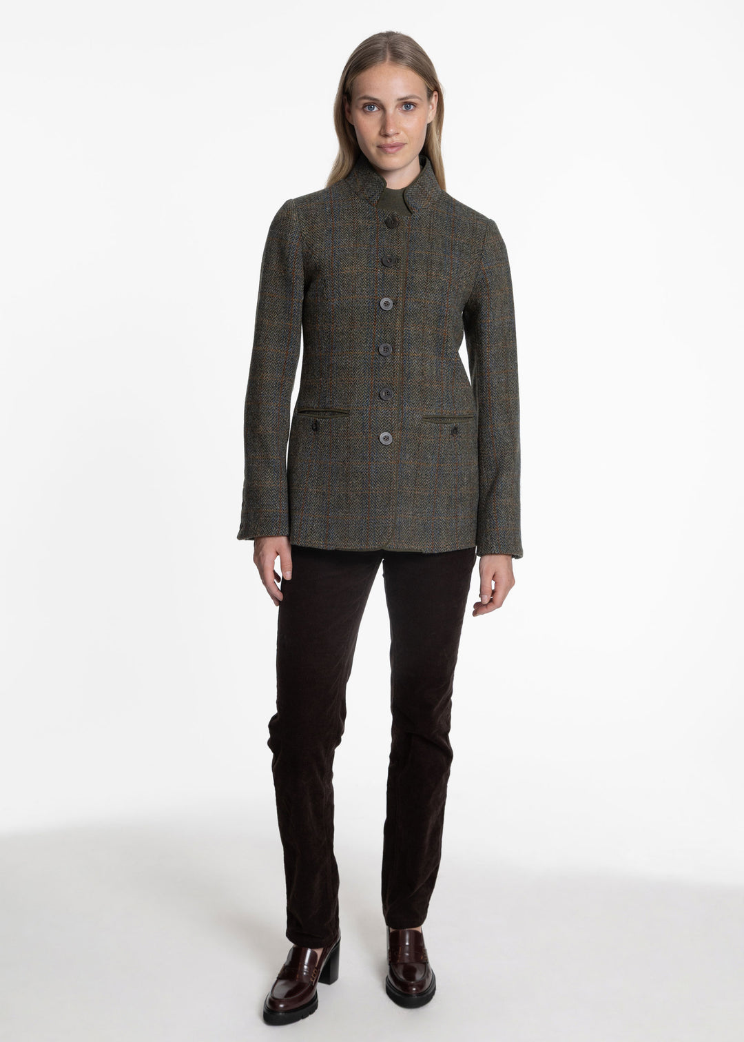 A woman modeling a dark olive green and brown tweed jacket with a high stand collar, button front, and flap pockets, paired with dark brown trousers and brown leather shoes, against a plain white studio background.