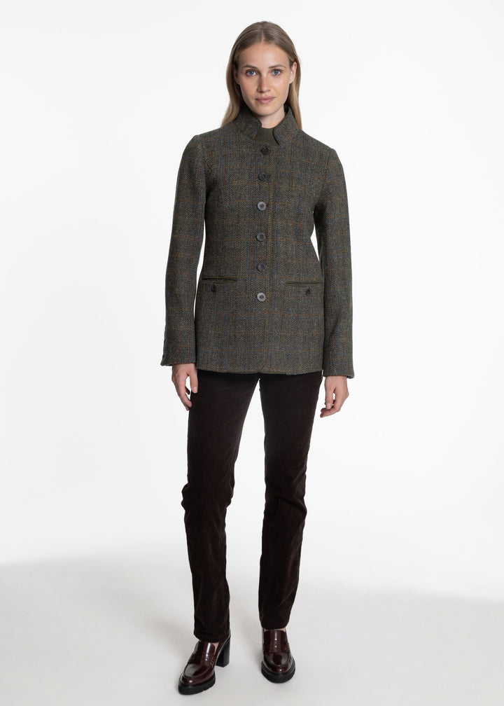 A woman modeling a dark olive green and brown tweed jacket with a high stand collar, button front, and flap pockets, paired with dark brown trousers and brown leather shoes, against a plain white studio background.