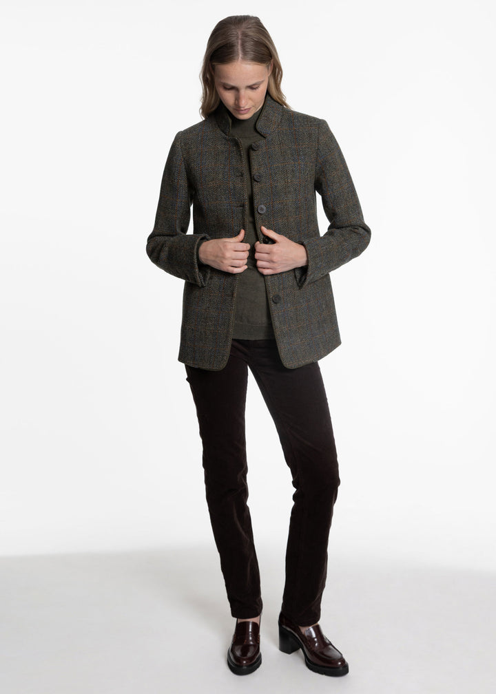 A woman modeling a dark green tweed jacket with a high collar and button front, paired with black trousers and brown leather shoes, against a plain white studio background.