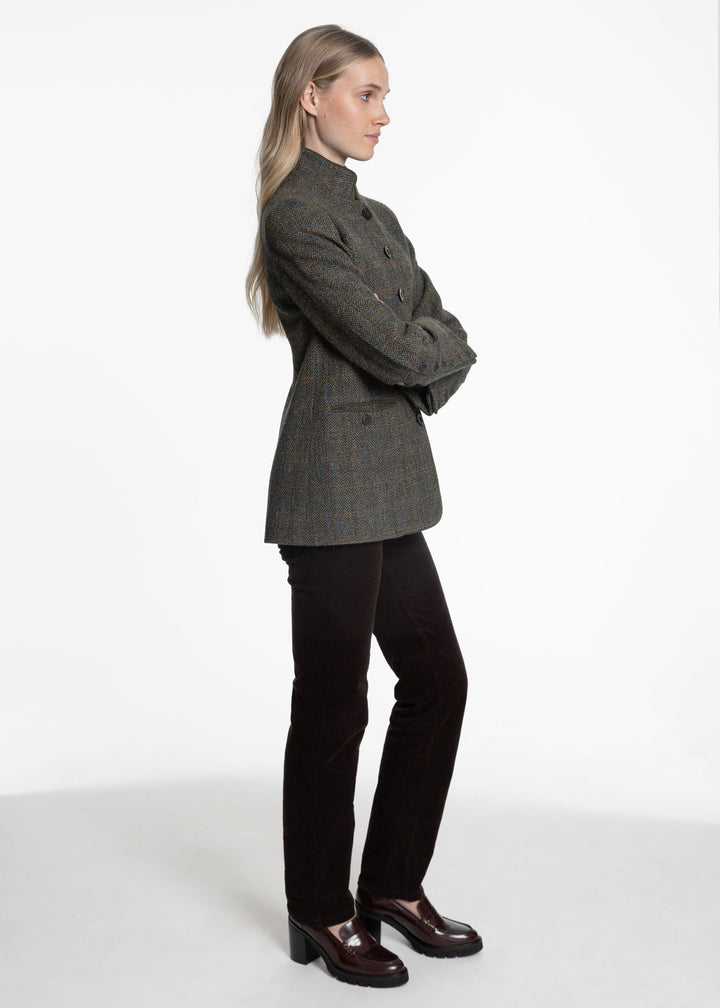 A woman stands in profile wearing a dark green tweed jacket with a high collar, black trousers, and dark brown leather shoes against a plain white studio background.