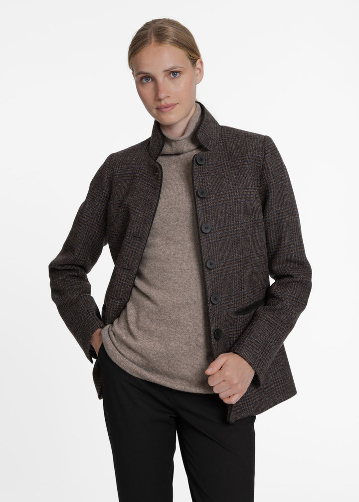 A woman modeling a dark brown and black checkered jacket over a taupe turtleneck sweater and black trousers, photographed against a clean white studio background.