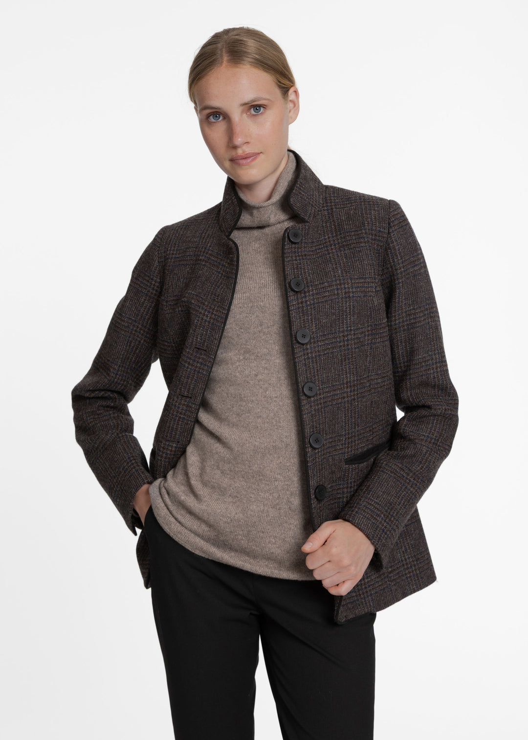 A woman modeling a dark brown and black checkered jacket over a taupe turtleneck sweater and black trousers, photographed against a clean white studio background.