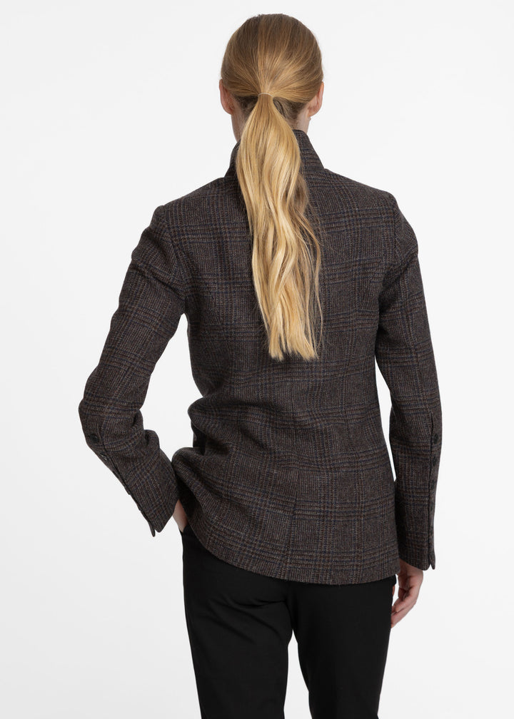 A woman shown from the back wearing a dark brown and black plaid blazer with black trousers, set against a plain white studio background.