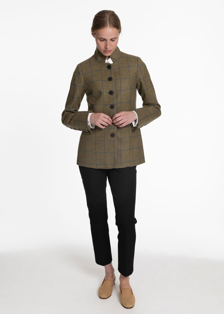 A woman modeling a green with blue plaid jacket with a high collar and black buttons, paired with black trousers and tan loafers, against a plain white studio background.