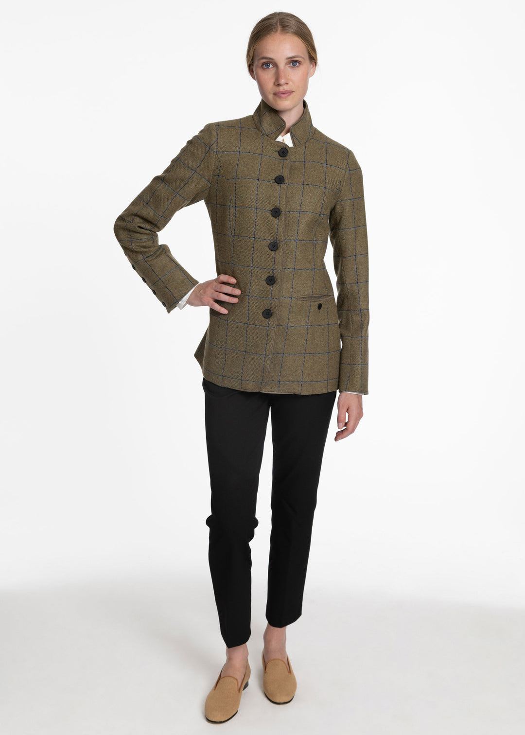 A woman modeling a green and blue plaid jacket with a mandarin collar and black buttons, paired with black pants and tan loafers, against a plain white studio background.
