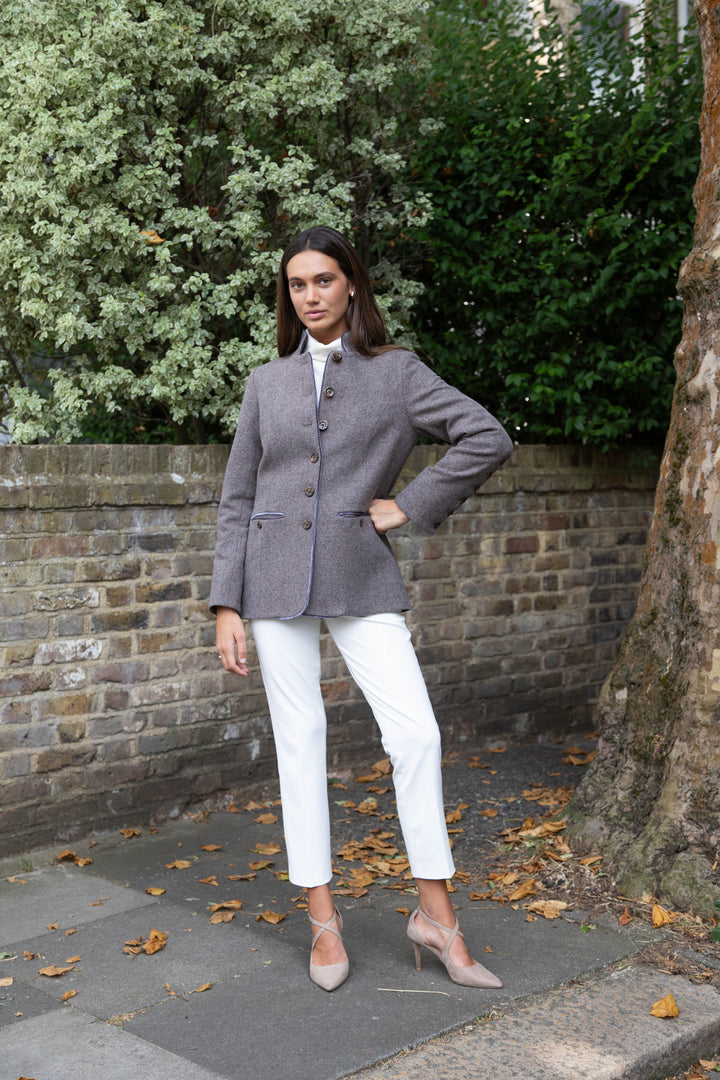 A woman stands outdoors on a sidewalk in front of a brick wall and trees, wearing a gray herringbone jacket with front buttons, purple velvet trim and white cropped pants, and nude pointed-toe heels.