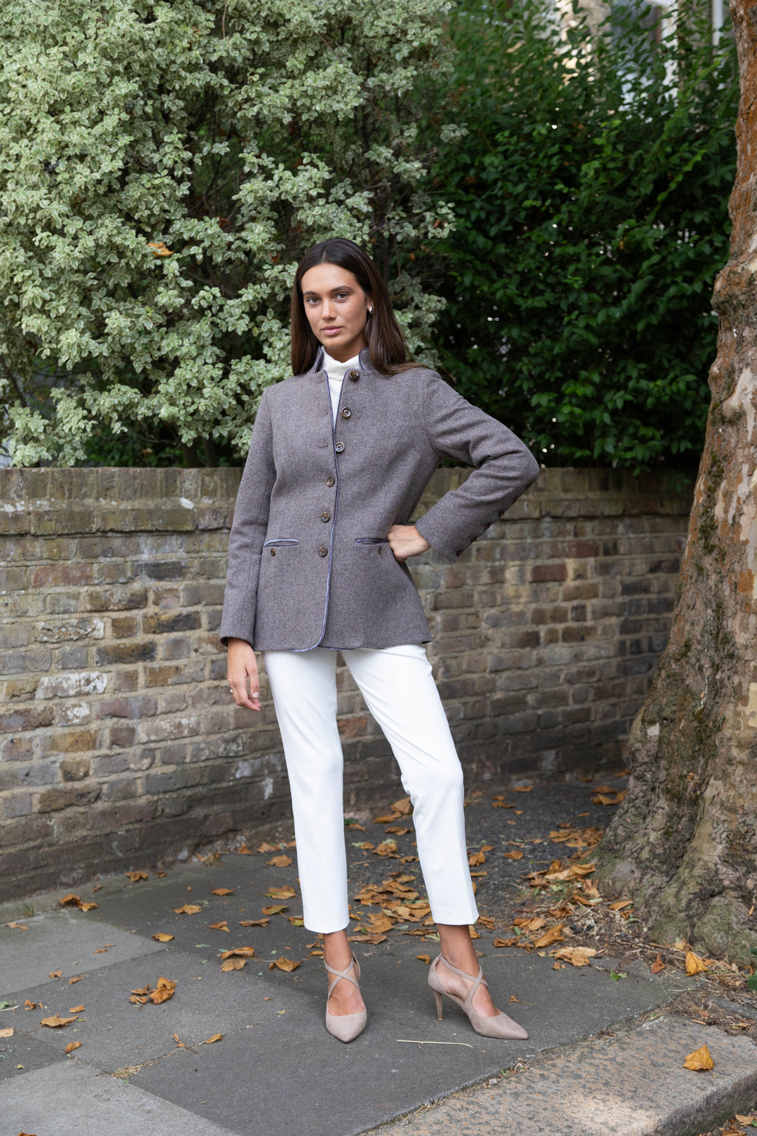 A woman stands outdoors on a sidewalk in front of a brick wall and trees, wearing a gray herringbone jacket with front buttons, purple velvet trim and white cropped pants, and nude pointed-toe heels.