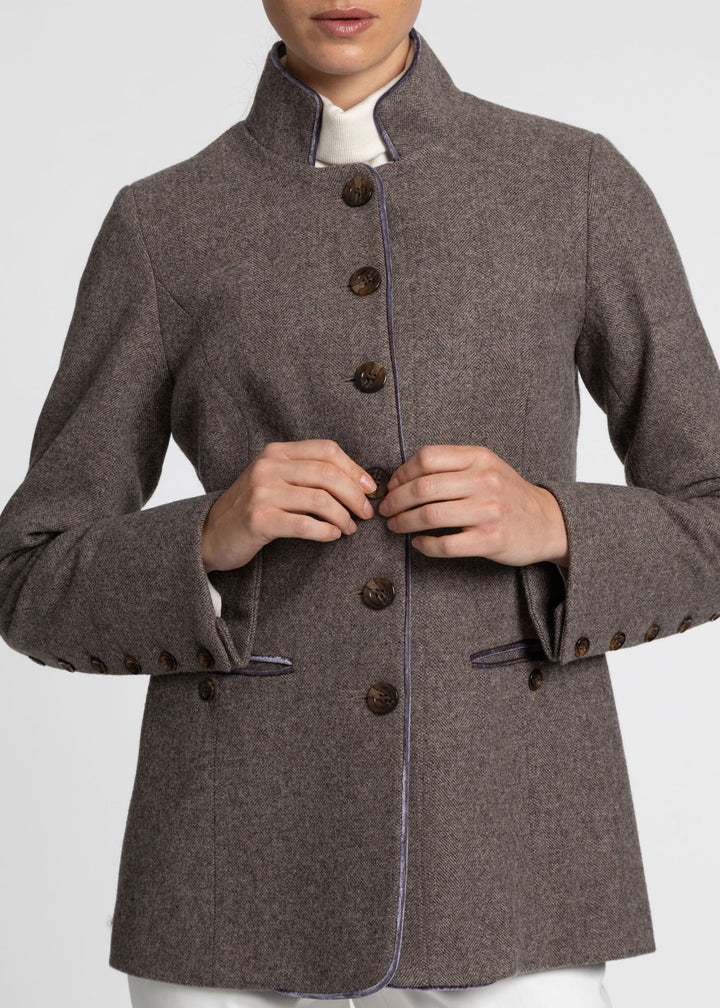 A close-up of a model wearing a brown-grey wool jacket with a high stand collar, dark brown buttons, and a light purple piping detail along the front and cuffs.