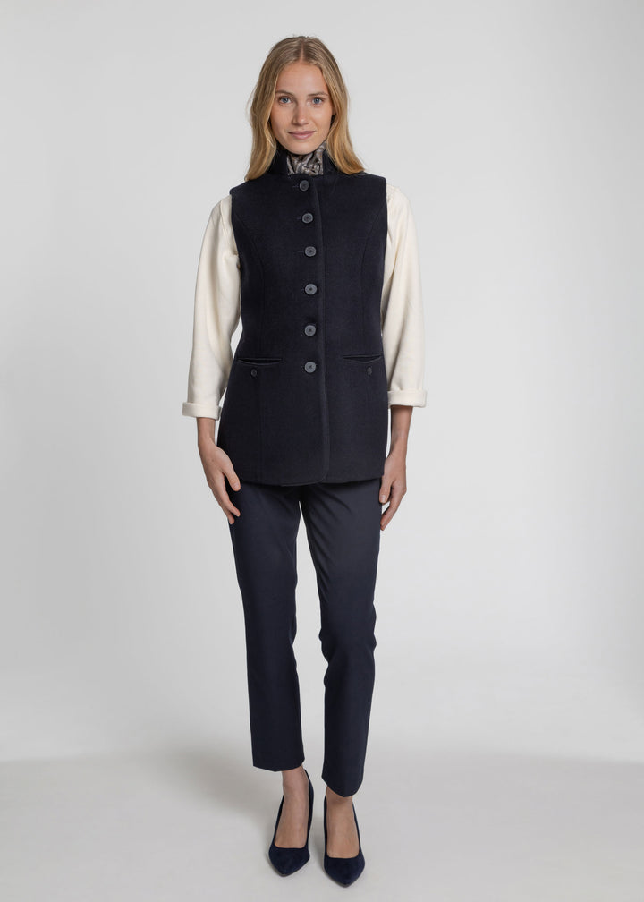 A woman stands against a light gray studio background wearing a dark navy sleeveless waistcoat over a cream long-sleeve blouse, matching dark navy trousers, and dark navy heels.