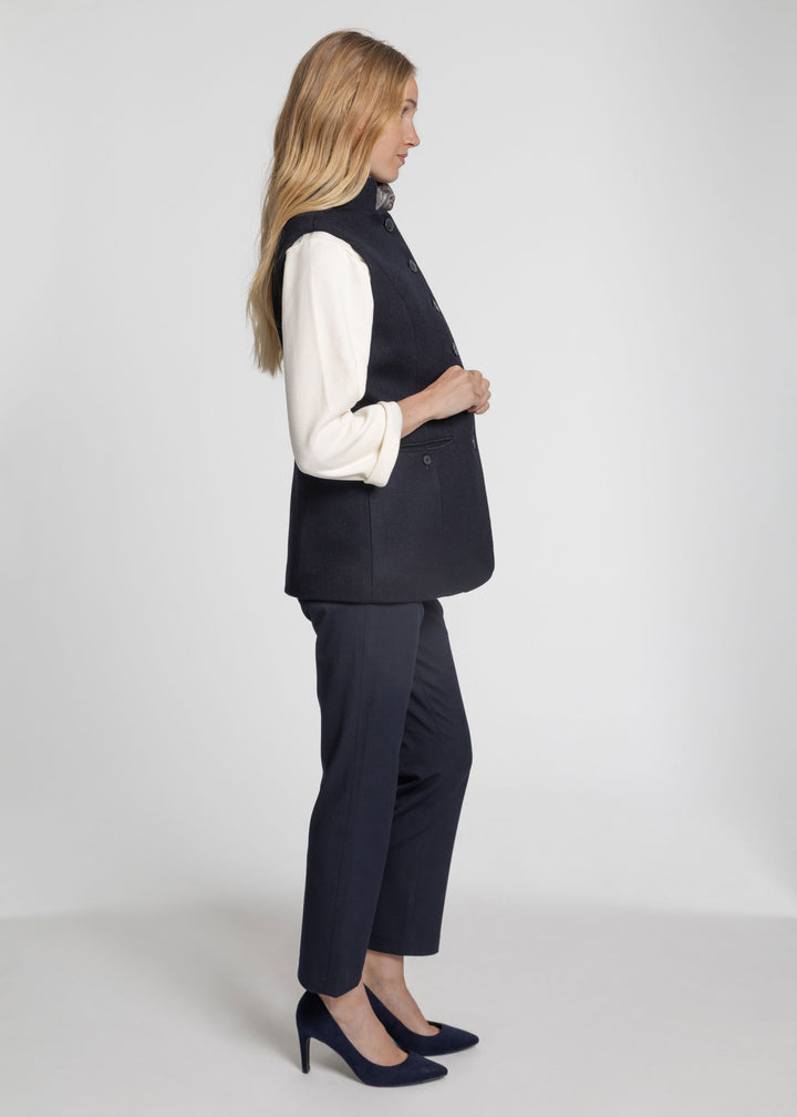 A woman stands in profile wearing a dark navy sleeveless waistcoat over a white long-sleeve blouse, matching navy trousers, and navy heels against a light gray studio background.