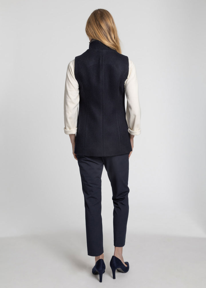 A woman shown from the back wearing a sleeveless dark navy waistcoat over a cream long-sleeve blouse, dark navy trousers, and navy heels against a light gray studio background.