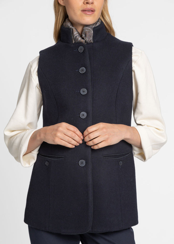 A woman wearing a dark navy sleeveless button-front waistcoat over a cream long-sleeve blouse, shown from the shoulders to the hips against a light gray studio background.