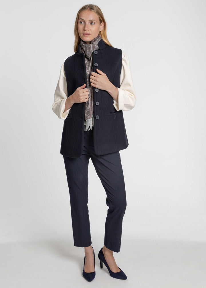 A woman modeling a dark navy sleeveless waistcoat over a cream long-sleeve blouse, paired with navy trousers, a gray patterned scarf, and navy heels, against a light gray studio background.