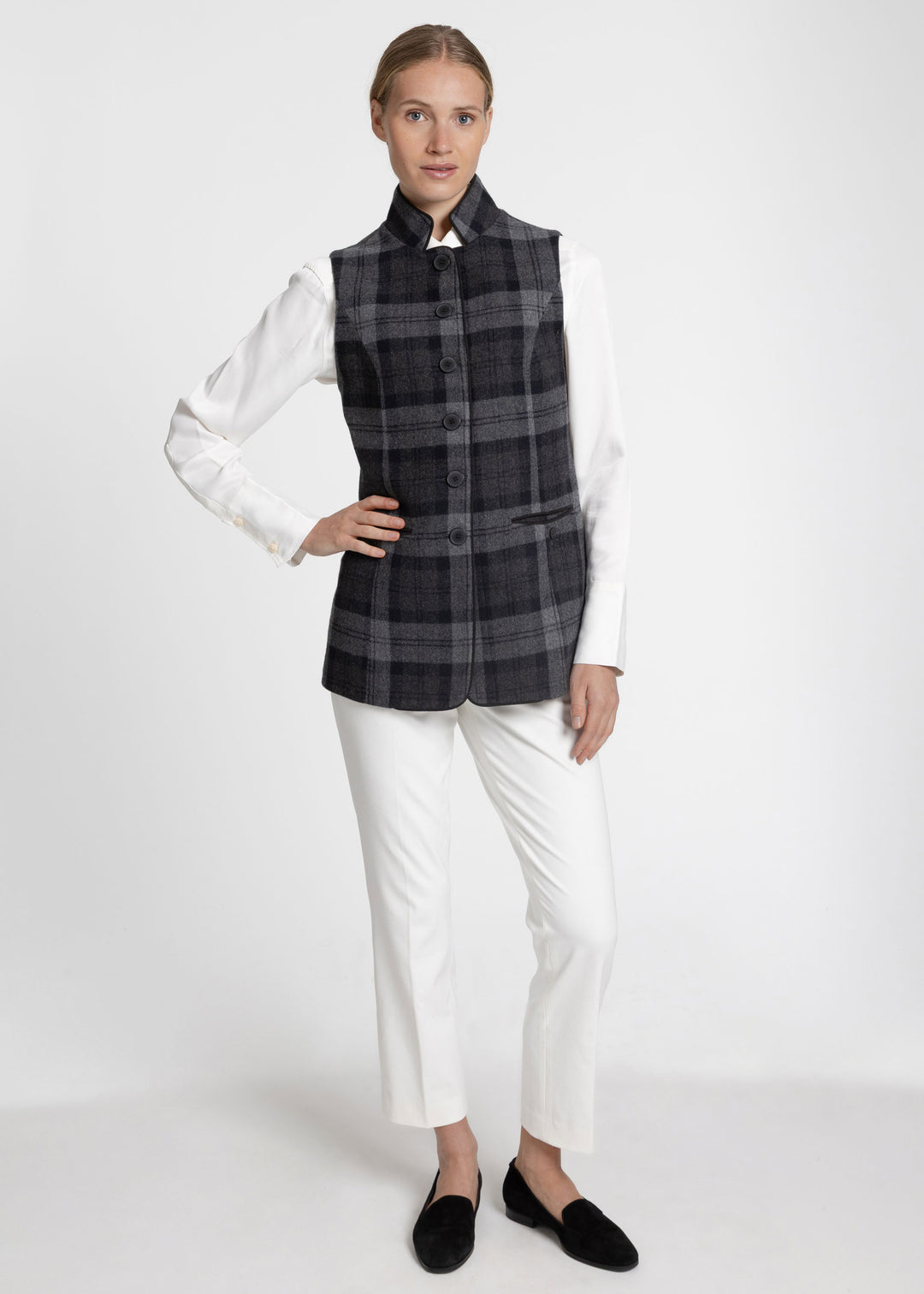 A woman wearing a grey and back plaid waistcoat over a white long-sleeve with white trousers. 