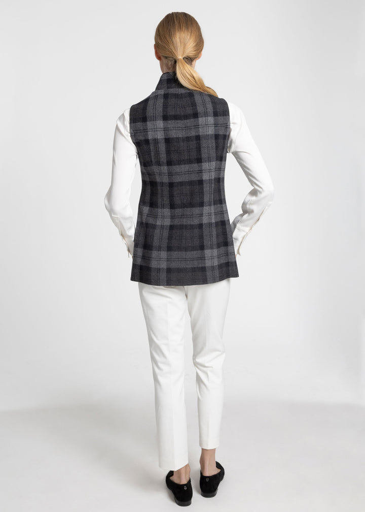 A woman shown from the back wearing a sleeveless black and gray plaid waistcoat over a white long-sleeve top and white pants, standing against a light gray studio background.