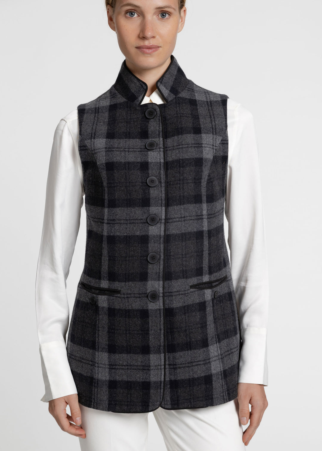 A woman wearing a sleeveless black and gray plaid waistcoat over a white long-sleeve shirt and white trousers, photographed against a light gray studio background.