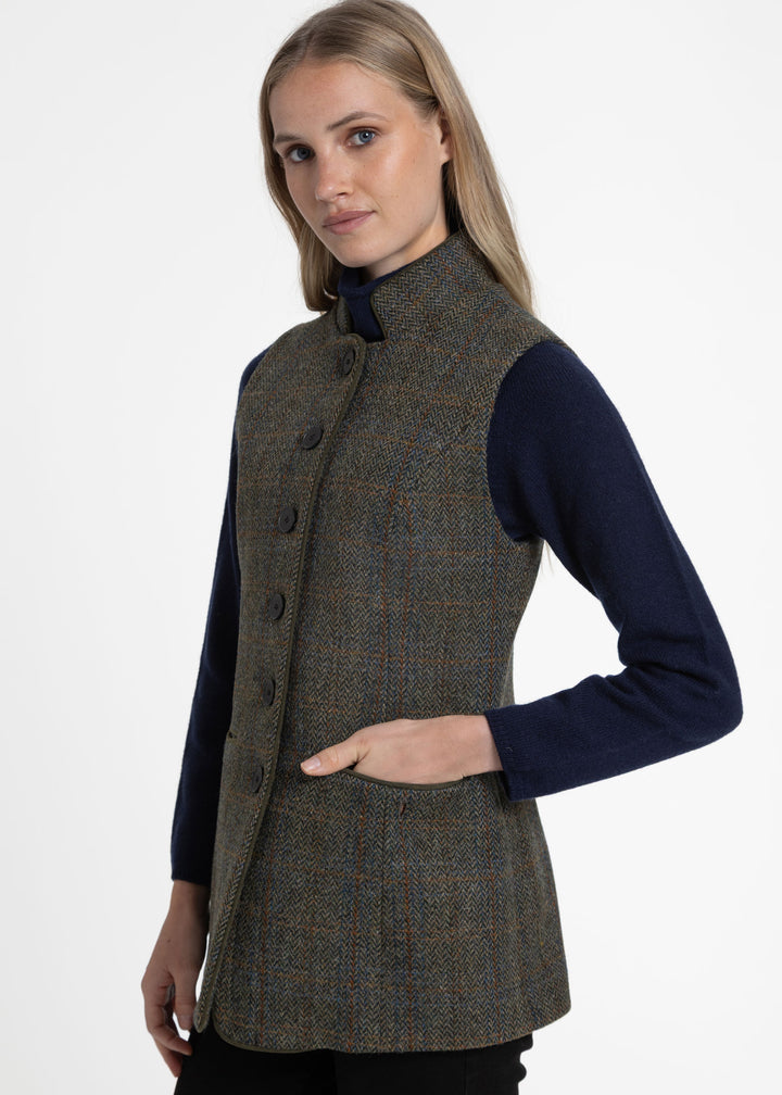 A woman wearing a sleeveless tweed waistcoat vest over a navy blue long-sleeve top, photographed against a plain light gray studio background.