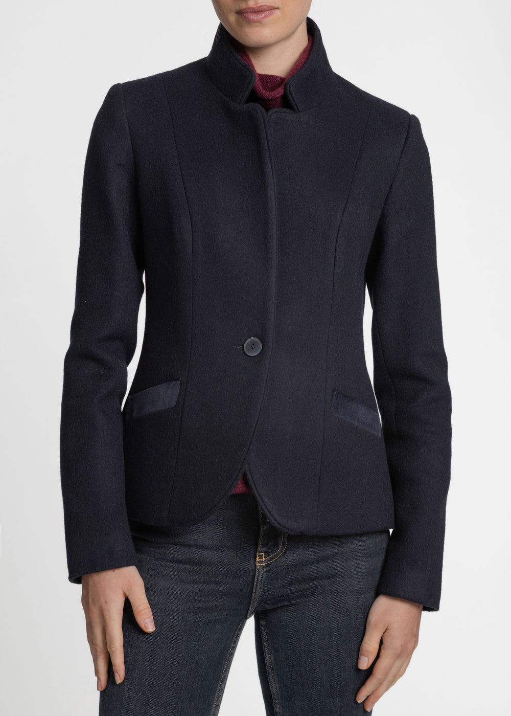 A woman wearing a dark navy blue cropped blazer with a stand collar, single button closure, and contrast pocket flaps, paired with dark jeans and a burgundy top, shown against a plain light gray background.