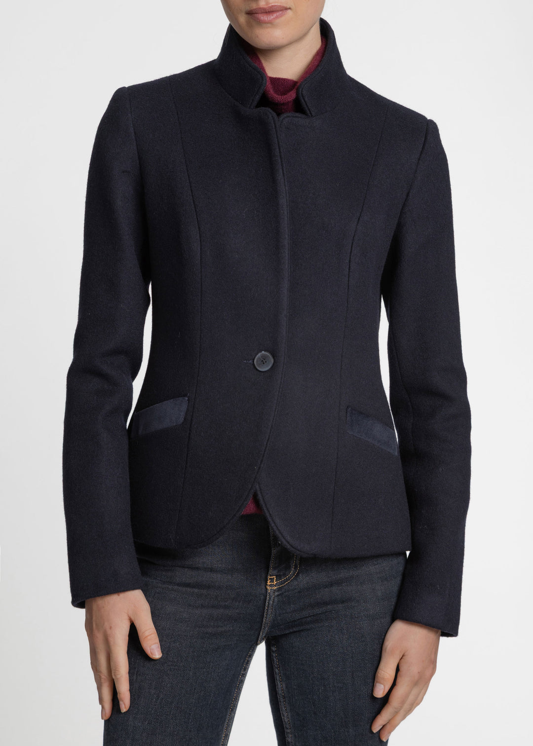 A woman wearing a dark navy blue cropped blazer with a stand collar, single button closure, and contrast pocket flaps, paired with dark jeans and a burgundy top, shown against a plain light gray background.