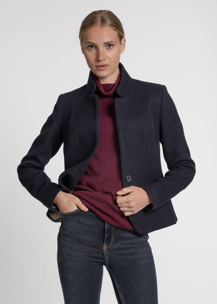 A woman modeling a dark navy blue structured jacket over a burgundy turtleneck sweater and dark gray jeans, photographed against a light gray studio background.