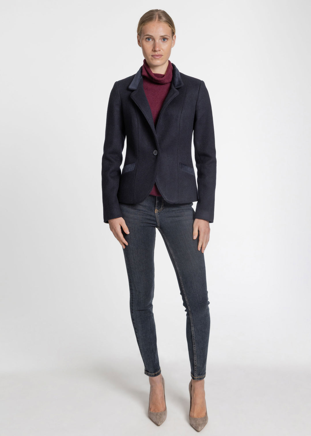A woman modeling a dark navy blue blazer over a burgundy turtleneck sweater, paired with dark gray skinny jeans and beige pointed-toe heels, against a plain white studio background.