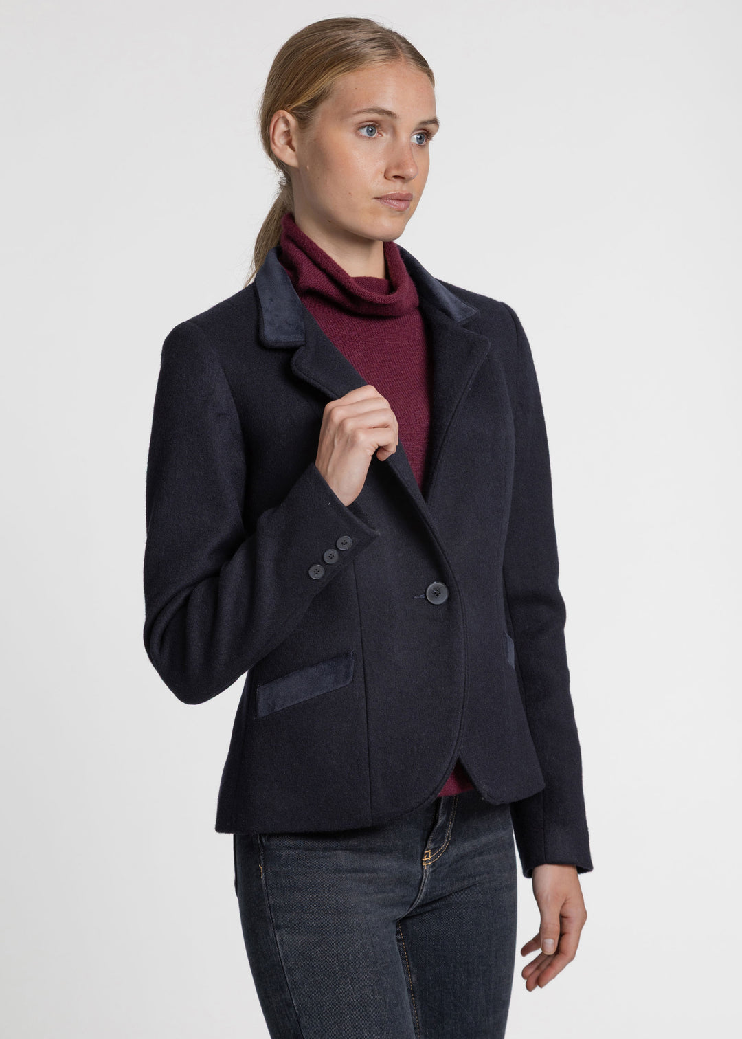 A woman modeling a dark navy wool-blend blazer with dark navy velvet lapels, worn over a burgundy turtleneck sweater and dark gray jeans, against a light gray studio background.