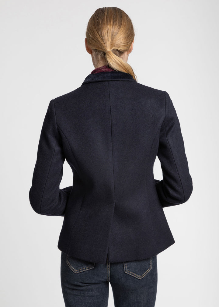 A woman viewed from the back wearing a dark navy tailored jacket with a notched collar and dark jeans, against a plain light gray studio background.