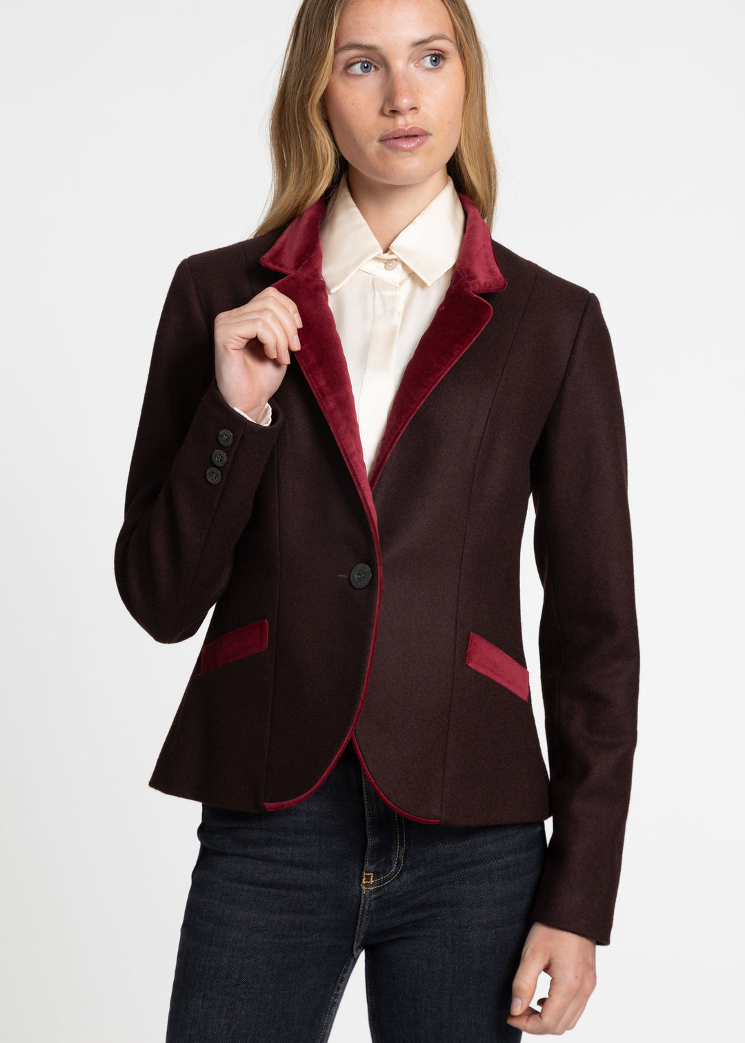 A woman modeling a dark mulberry blazer with a contrasting deep red velvet collar and pocket trim, worn over a cream blouse and dark blue jeans against a plain white background.
