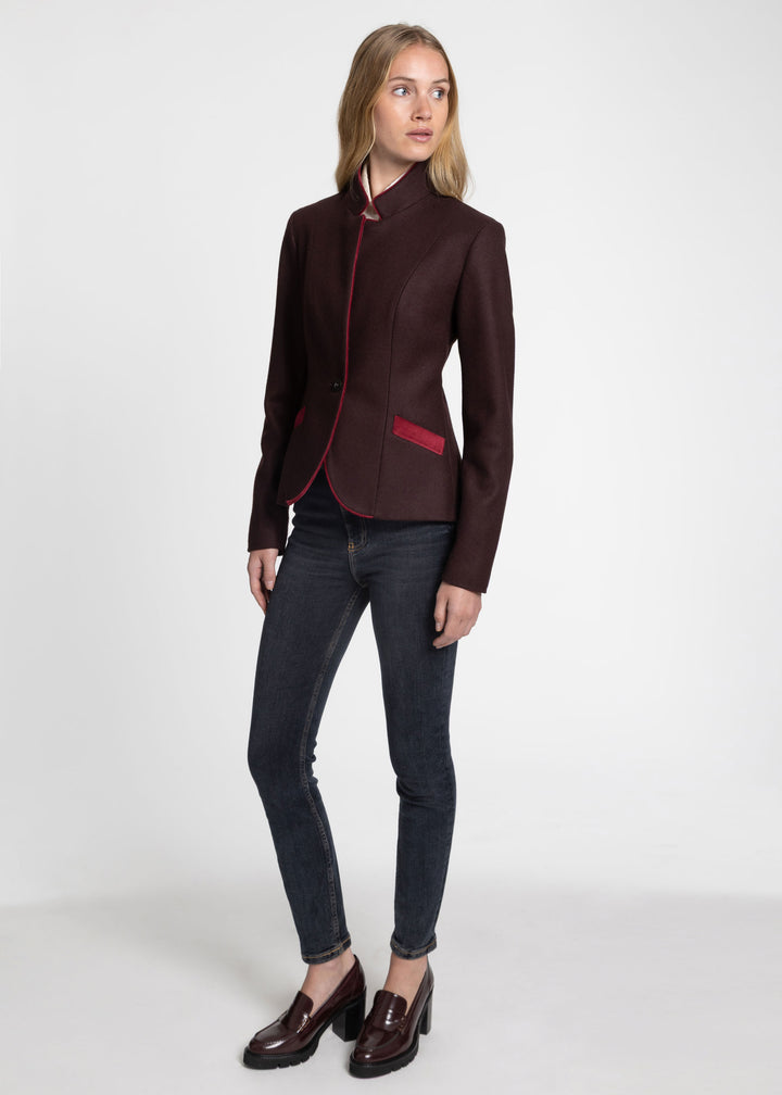 A woman modeling a dark mulberry structured blazer with red piping and a red pocket detail, paired with dark gray skinny jeans and brown leather loafers, against a plain white studio background.