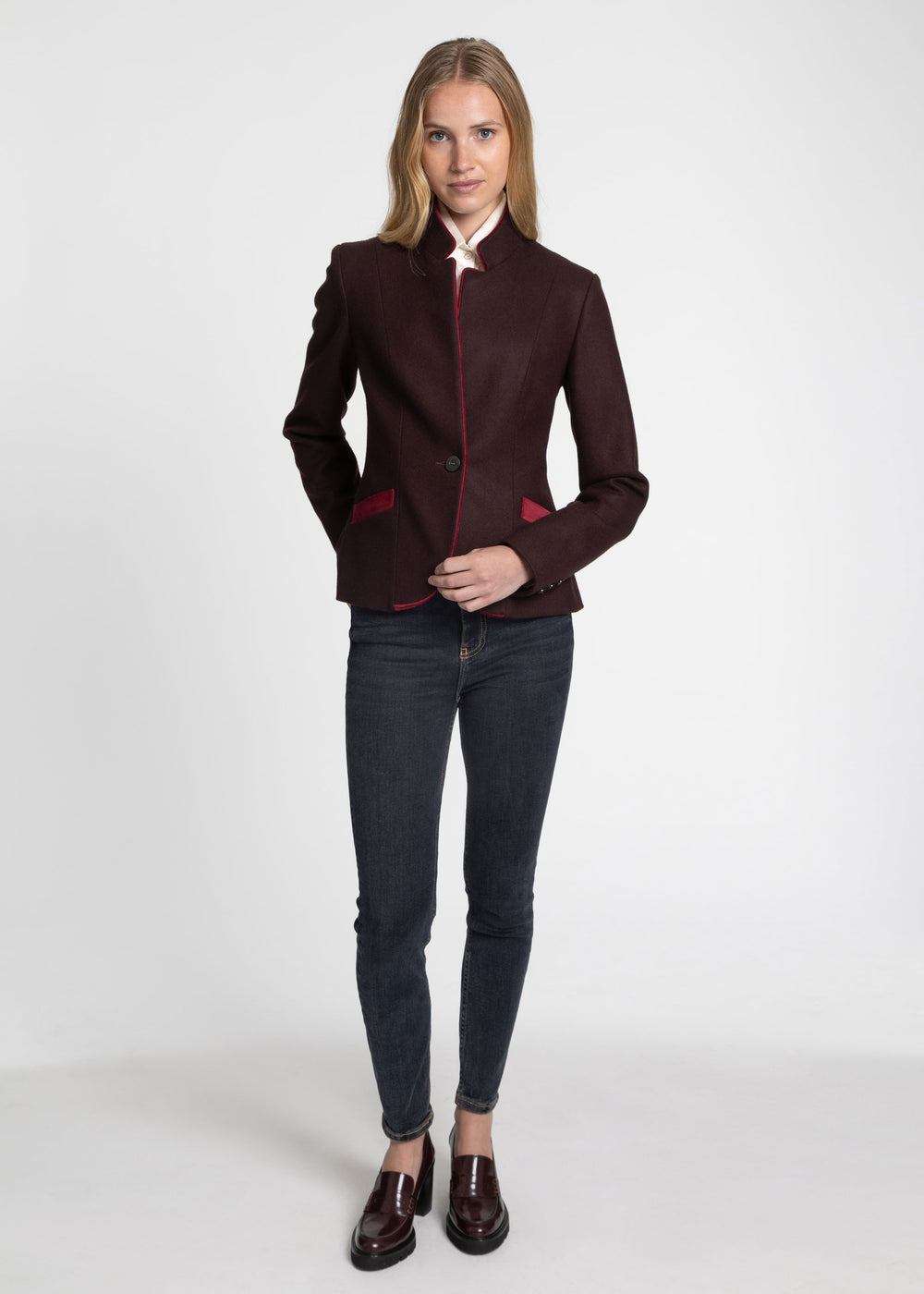 A woman modeling a dark burgundy wool-blend blazer with contrasting red trim on the collar, and pocket flaps, paired with dark gray skinny jeans and brown leather loafers, against a plain white studio background.