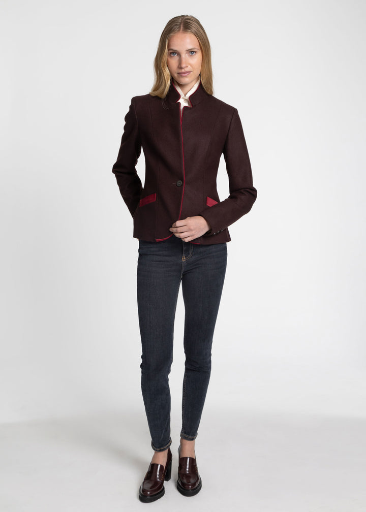 A woman modeling a dark burgundy wool-blend blazer with contrasting red trim on the collar, and pocket flaps, paired with dark gray skinny jeans and brown leather loafers, against a plain white studio background.