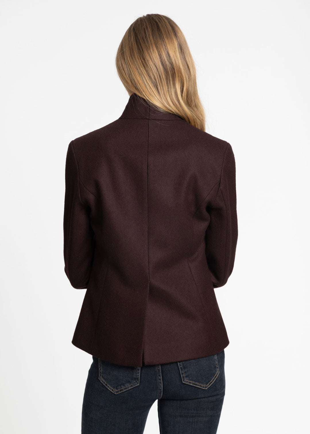 A woman viewed from the back wearing a dark mulberry structured blazer and dark blue jeans against a plain white background.