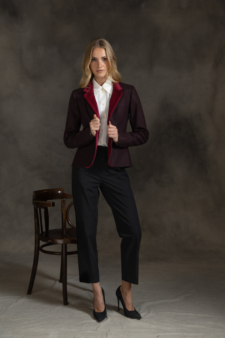 A woman models a tailored blazer in deep burgundy with bright red velvet lapels, paired with black trousers, a white collared shirt, and black heels, standing in front of a dark textured studio backdrop next to a wooden chair.