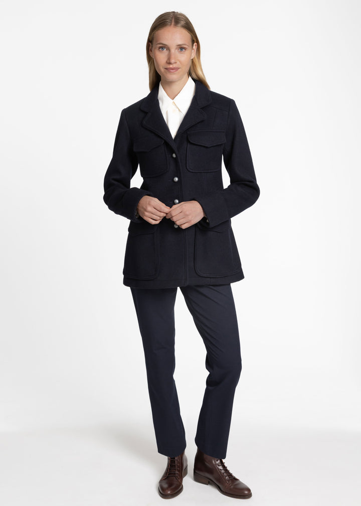 A woman modeling a dark navy blue tailored jacket with flap pockets, worn over a white collared shirt and paired with matching navy trousers and brown leather shoes, against a plain white studio background.