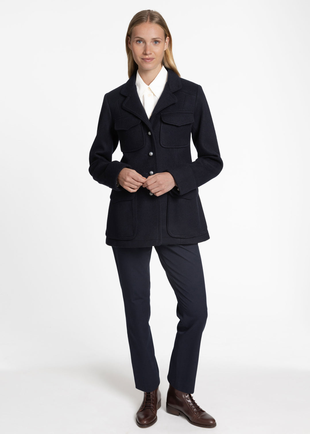 A woman modeling a dark navy blue tailored jacket with flap pockets, worn over a white collared shirt and paired with matching navy trousers and brown leather shoes, against a plain white studio background.